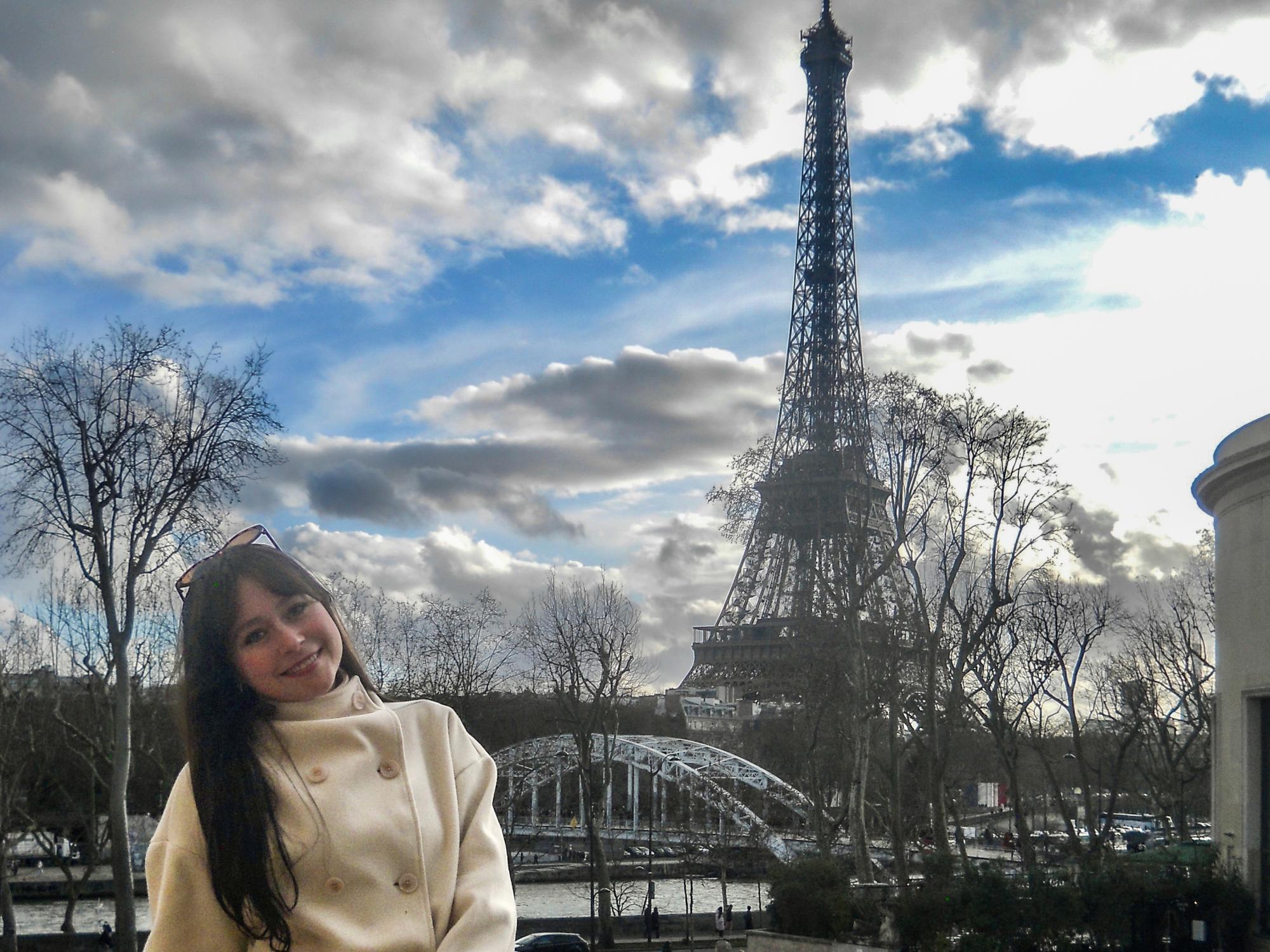 Jenna Guarrera wears a white jacket while posing in front of the Eiffel Tower.
