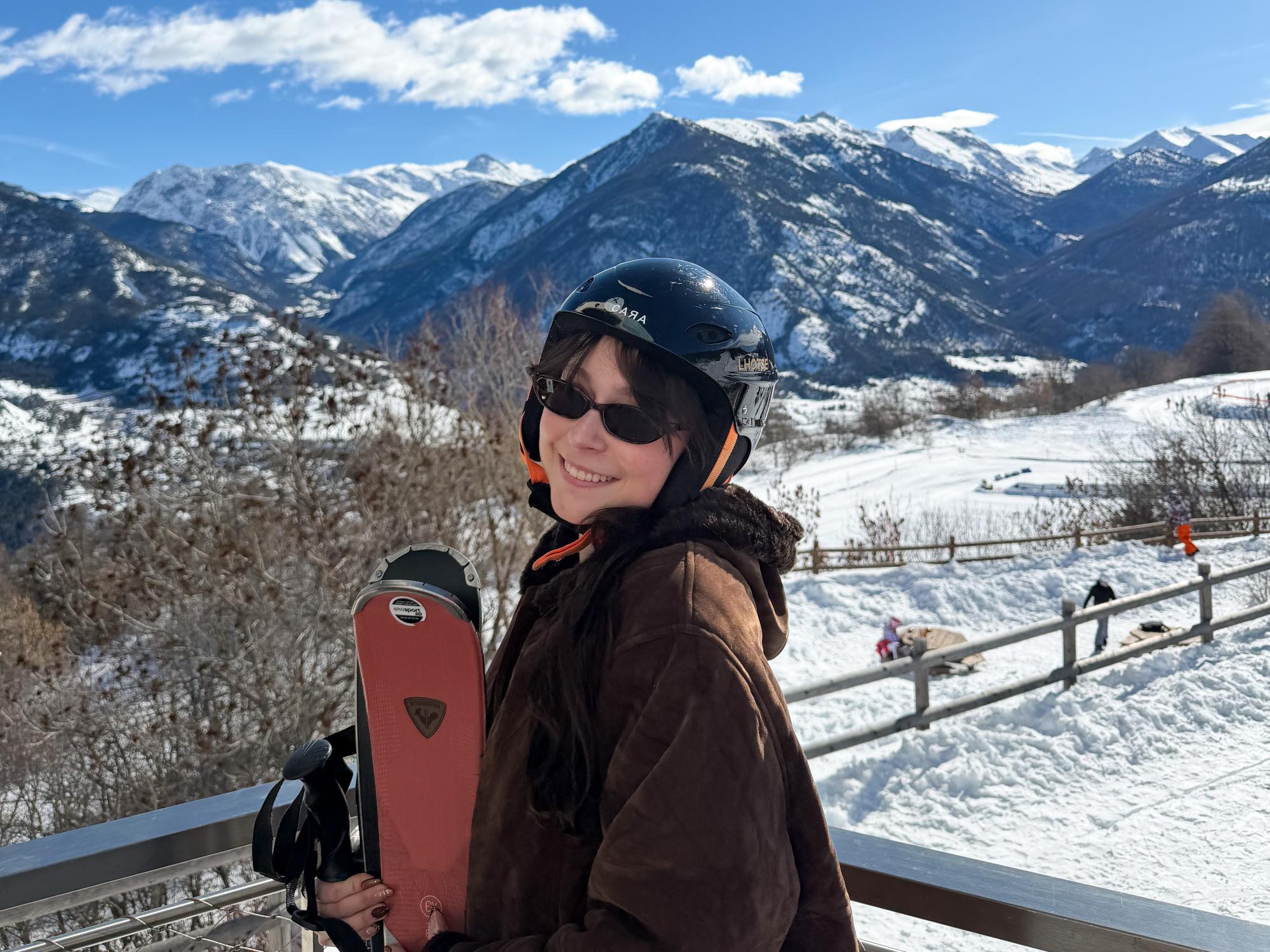 Jenna Guarrera wears a helmet and sunglasses while holding skis with the French Alps behind her.