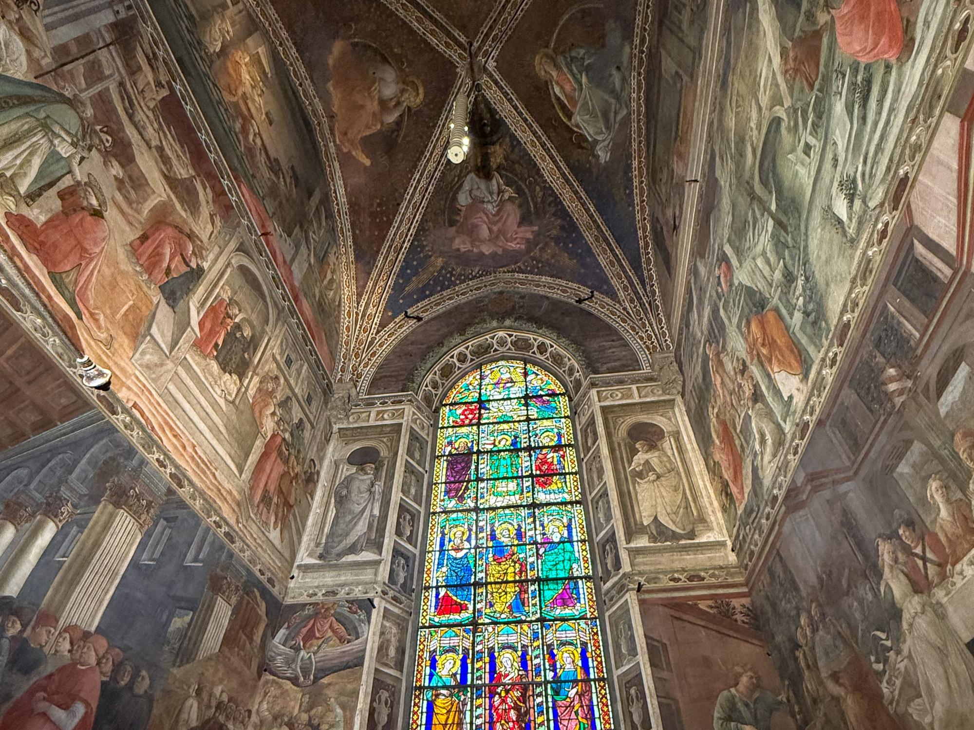 The interior walls of a chapel are covered in Renaissance frescoes with a tall arched stained-glass window at the center.