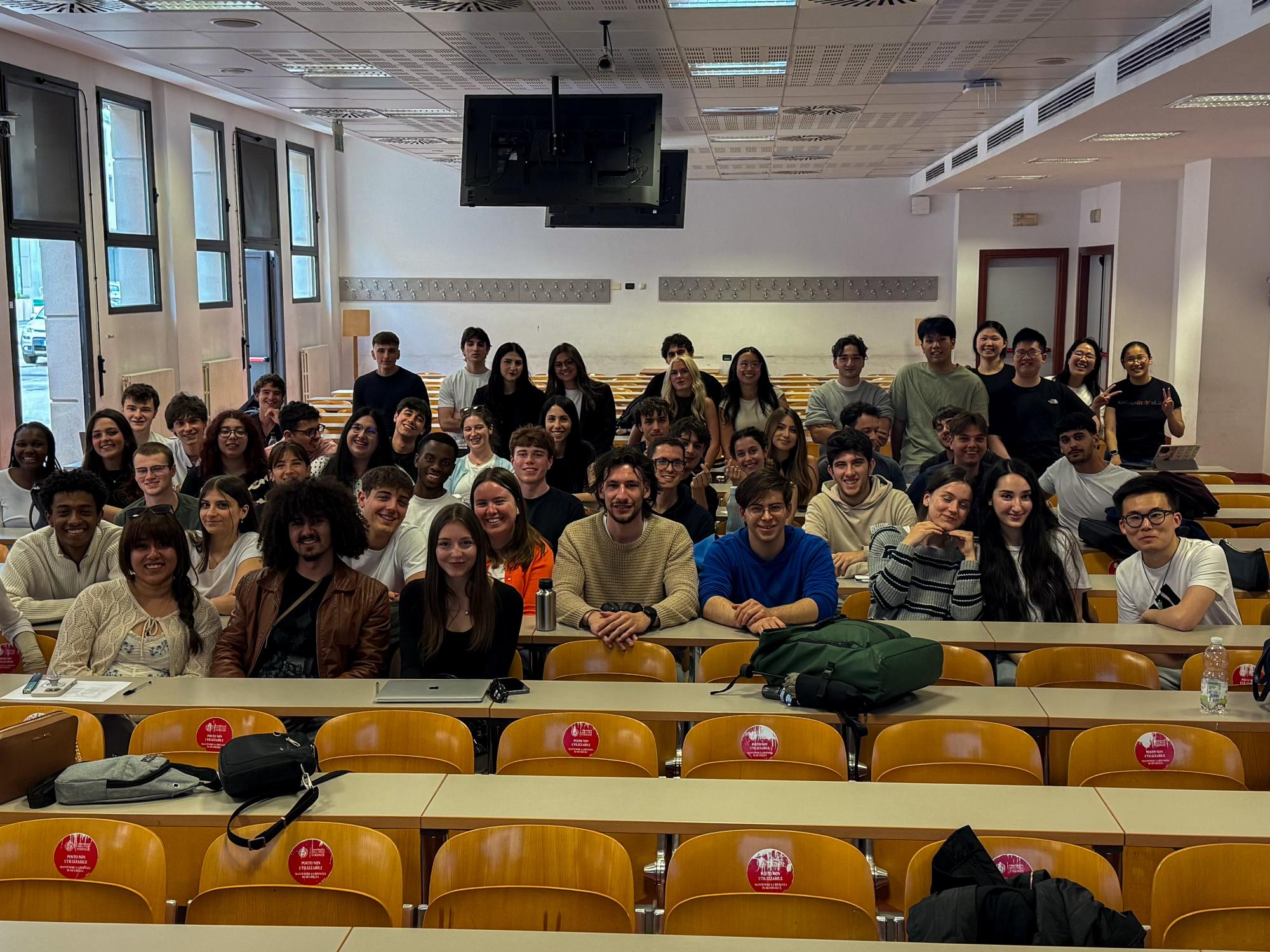 Fifty-three students smile for a group photo inside a bright university hall with wooden chairs and long rectangular tables.