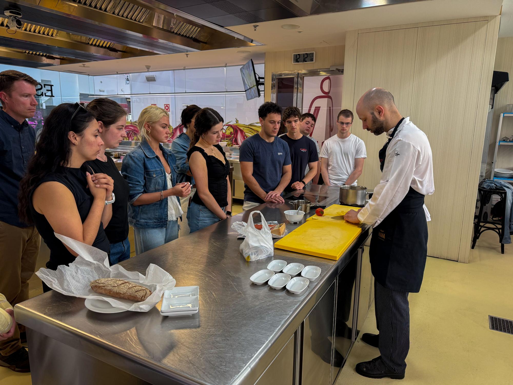 A chef in a white coat and black apron slices a tomato while about 10 students watch closely in a commercial kitchen.