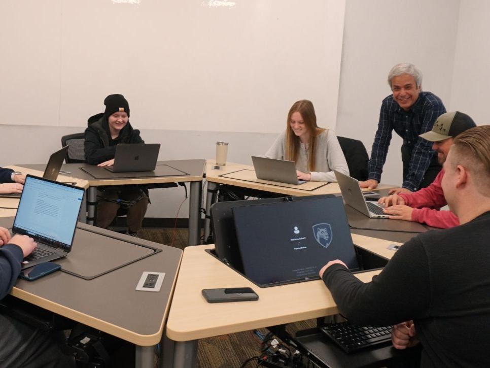 a group of students sit at a computer station discussing their project with their professor, who is standing next to the group and interacting with them