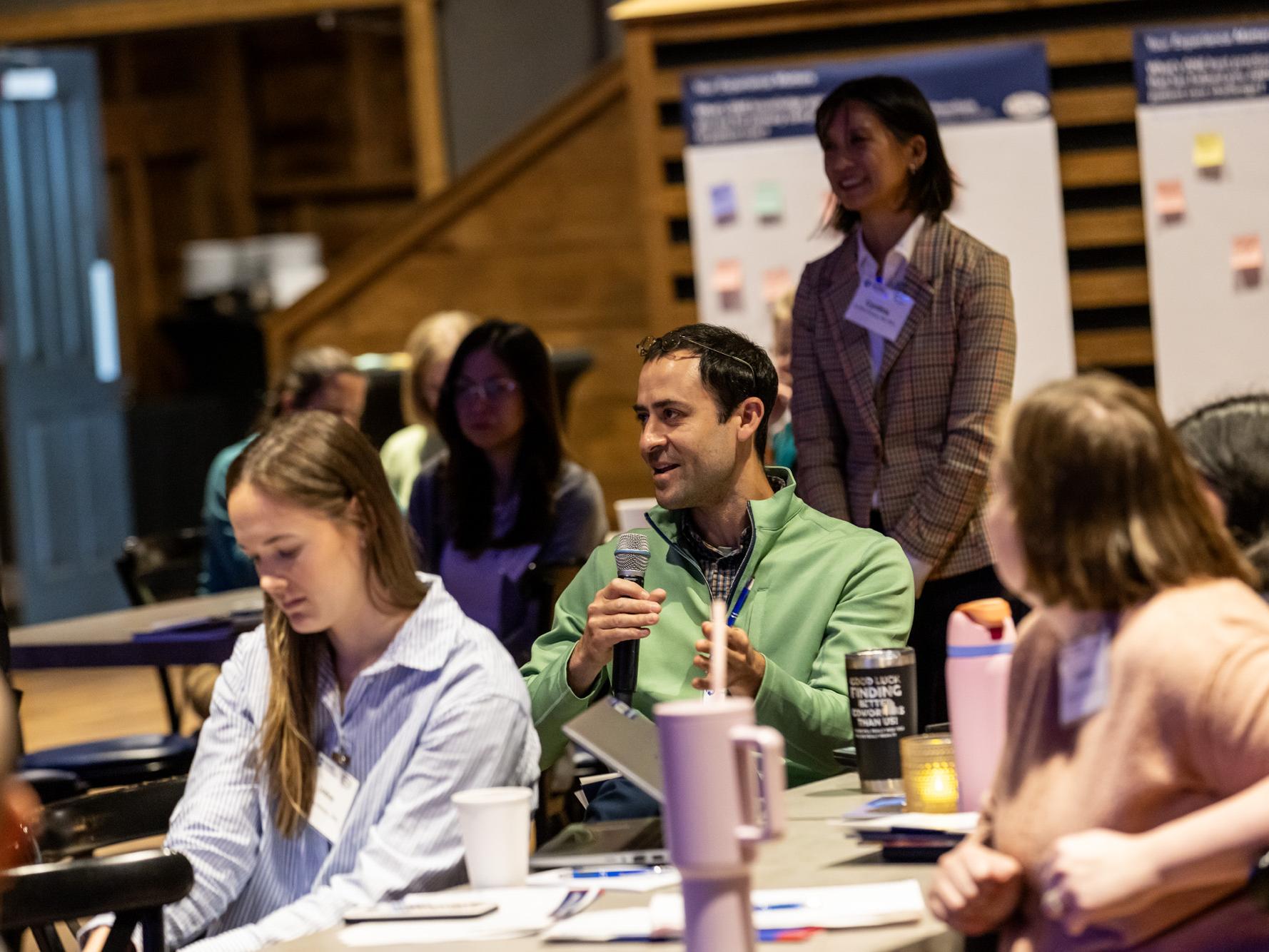 A male conference attendee in a green quarter-zip sweater speaks into a microphone while sitting at a table with other attendees.