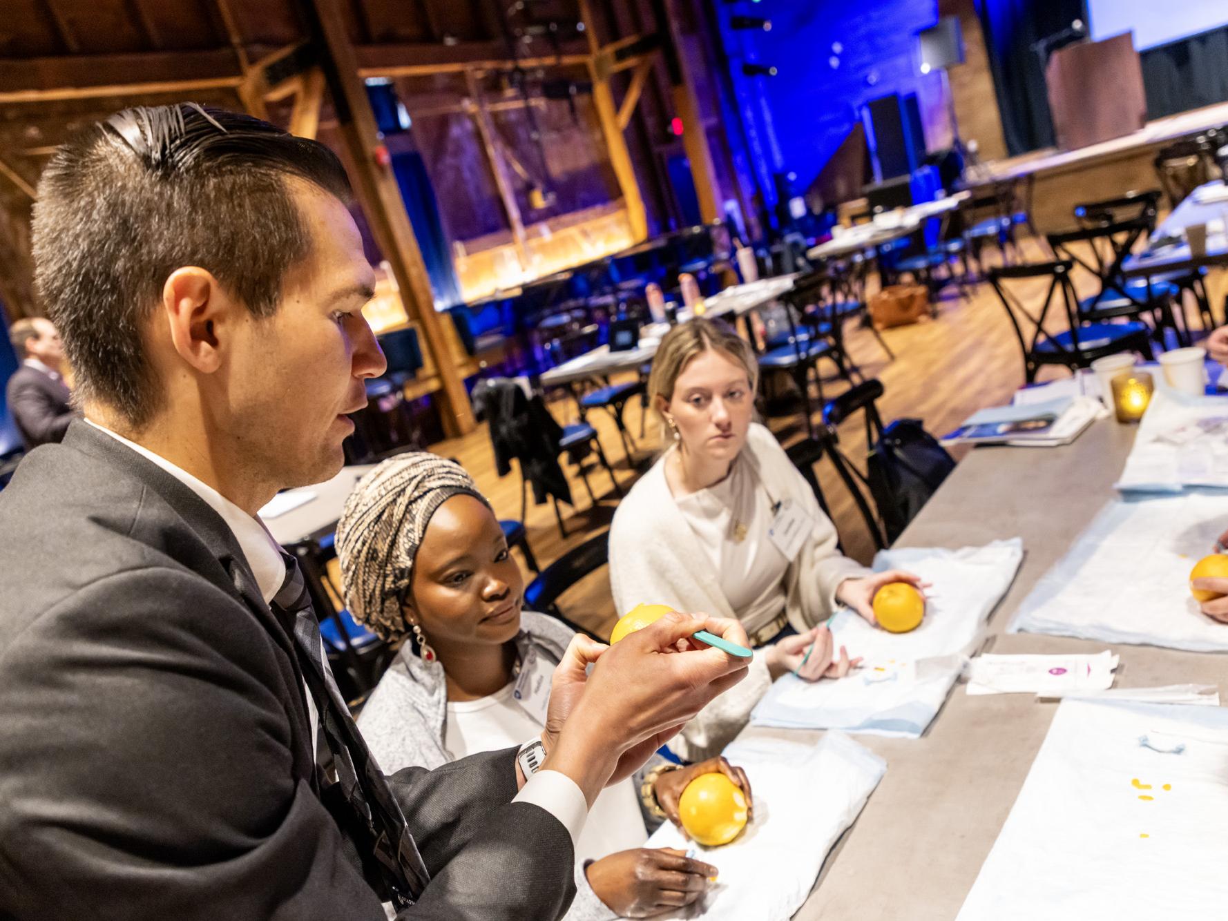 A male medical instructor in a suit demonstrates a medical procedure on an orange using a small tool while three female attendees watch closely at a conference table.
