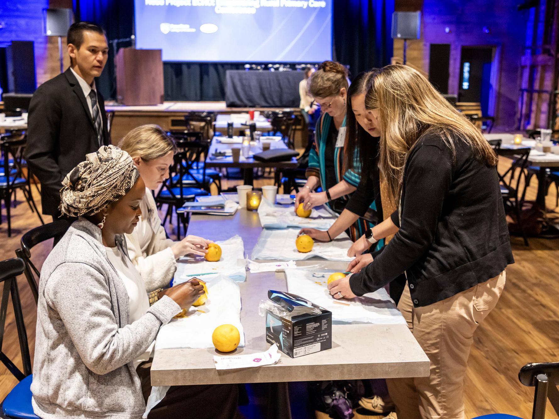 Four female medical professionals stand around a table practicing medical biopsy procedures on oranges laid out on medical pads.