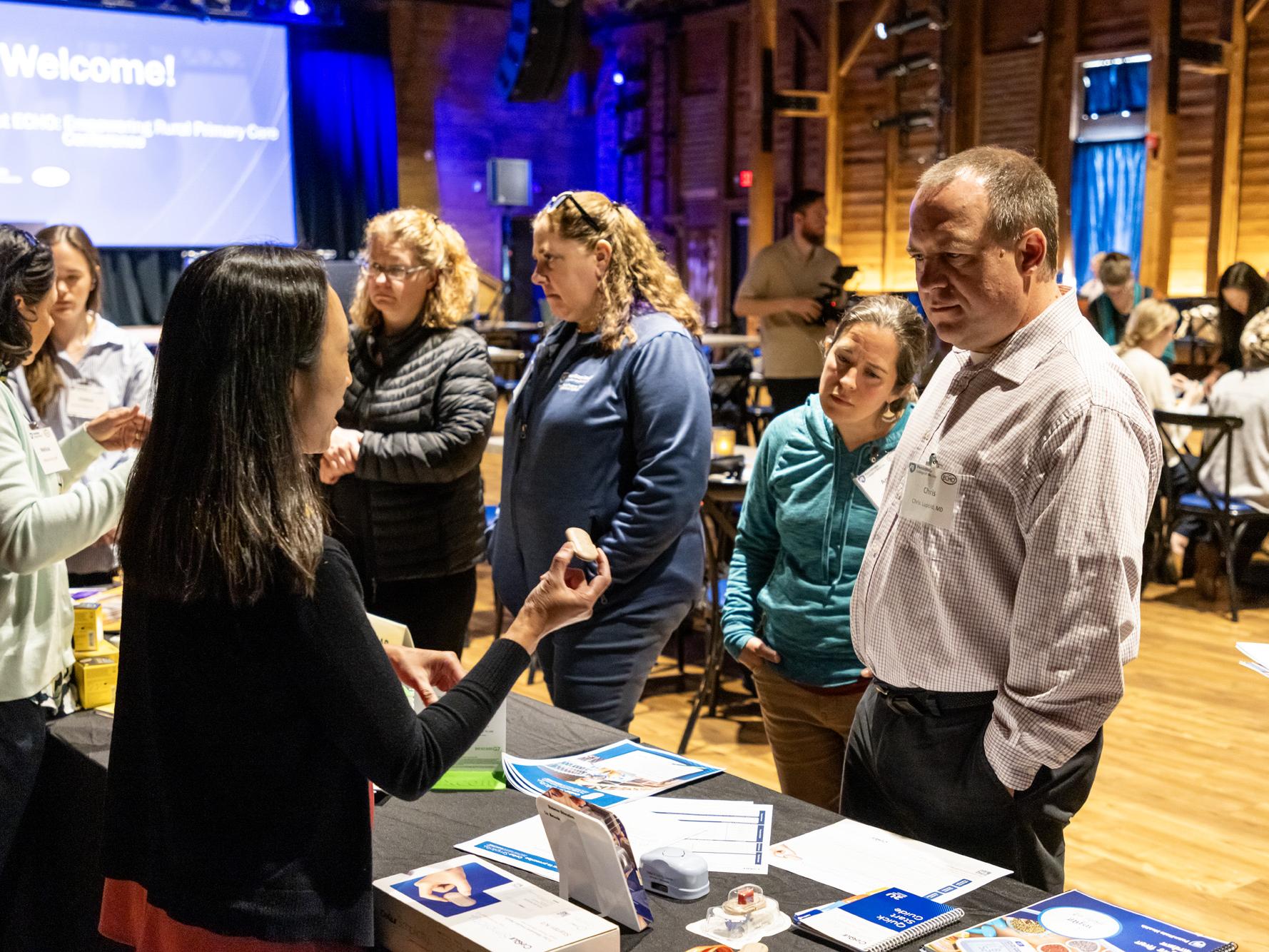  A female instructor holds up a small round, continuous glucose monitor device while explaining it to a man and a woman standing at a display table.