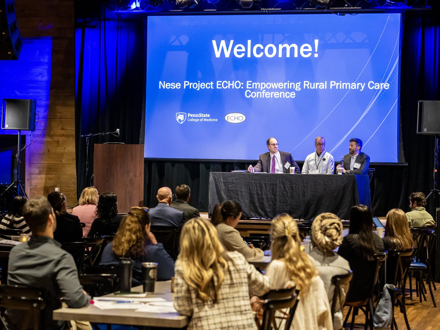 A wide shot of a conference room with attendees sitting at tables facing a brightly lit stage with a blue screen that reads "Welcome! Nese Project ECHO: Empowering Rural Primary Care Conference."