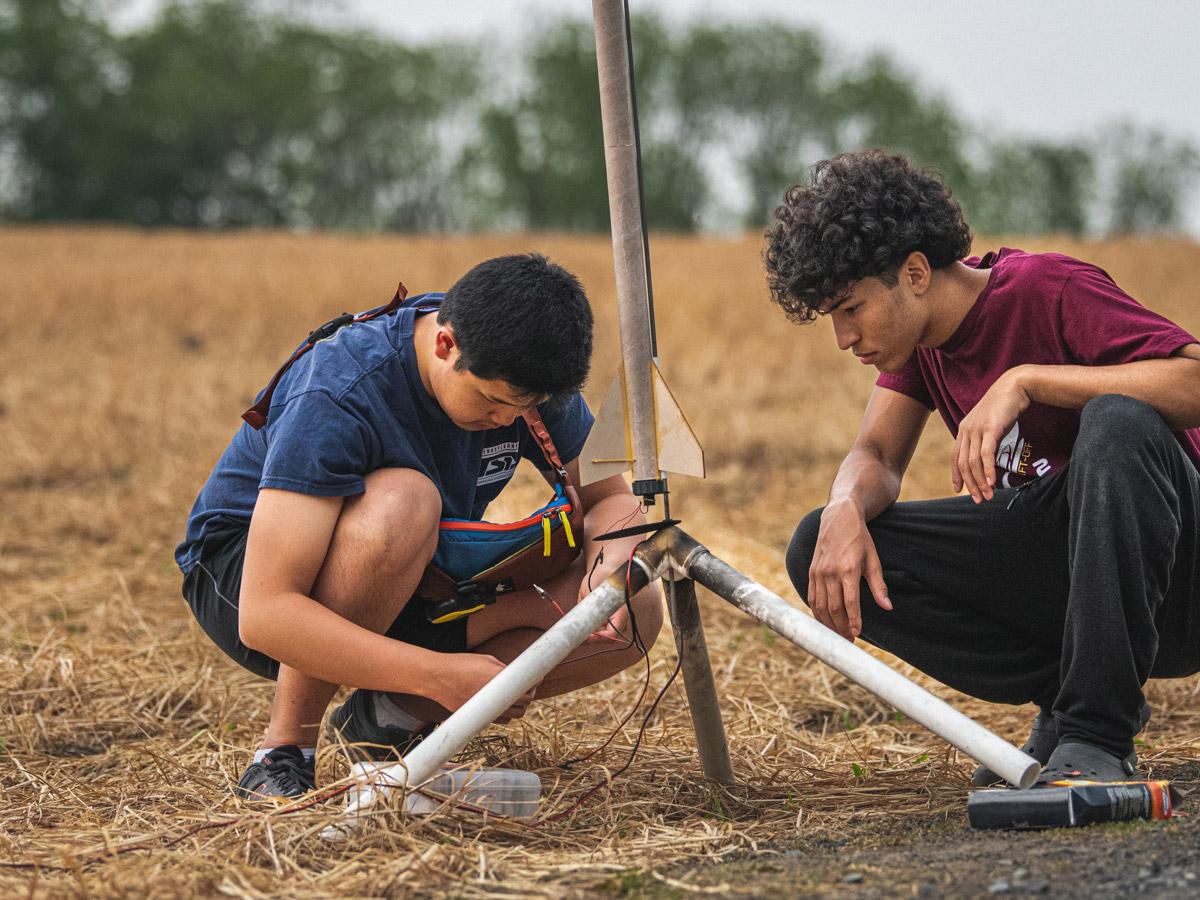 two students crouch on ground next to model rocket