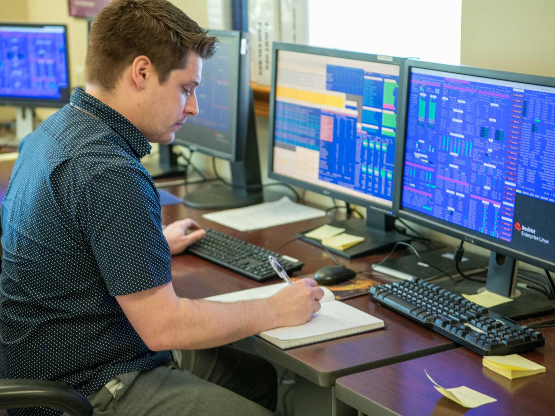 Tyler Sprague writes in notebook, with several computers displaying data on desk