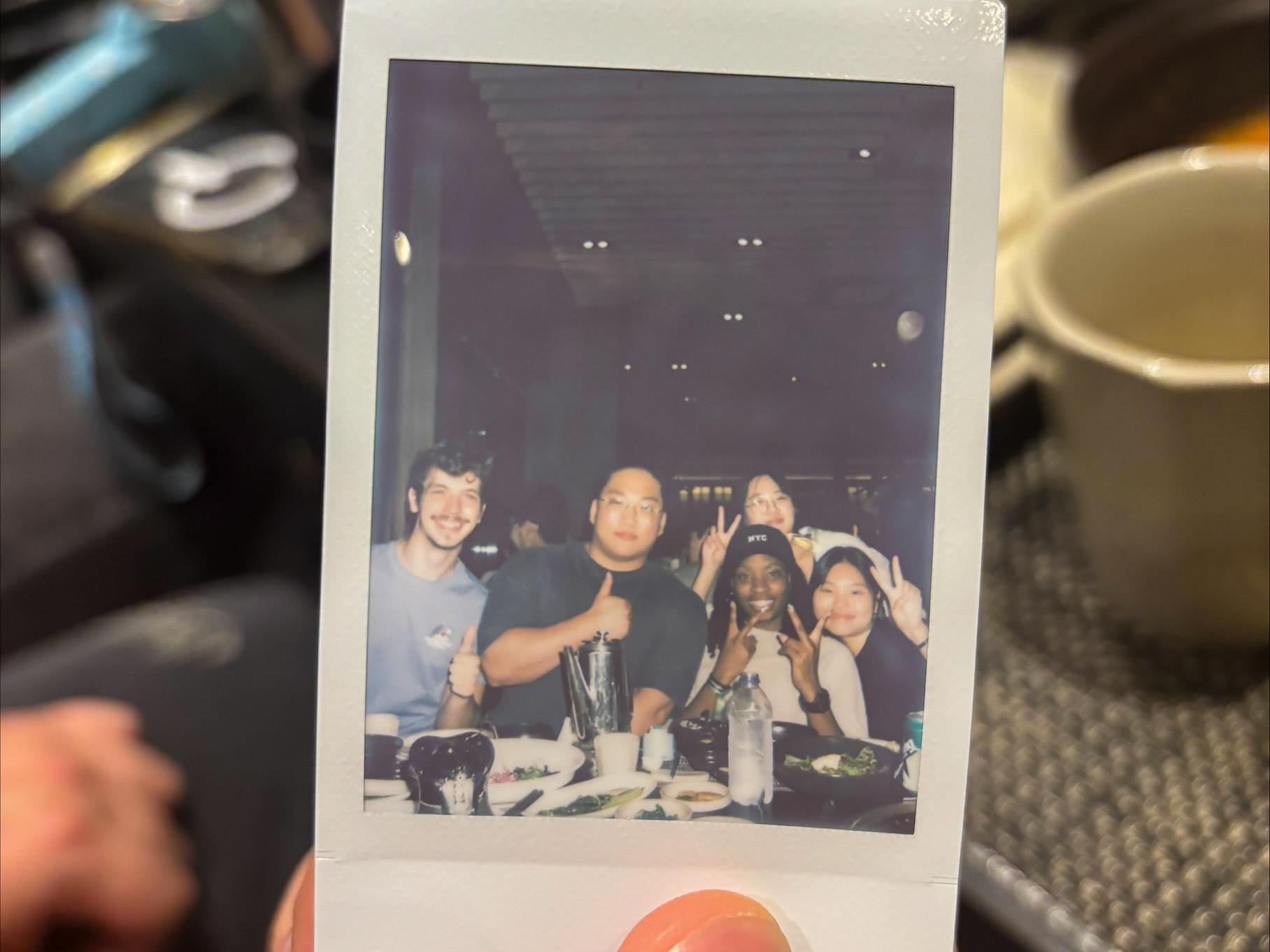 A hand holds a Polaroid photo showing five people seated at a restaurant table, smiling and making peace signs over plates, bowls, and drinks.