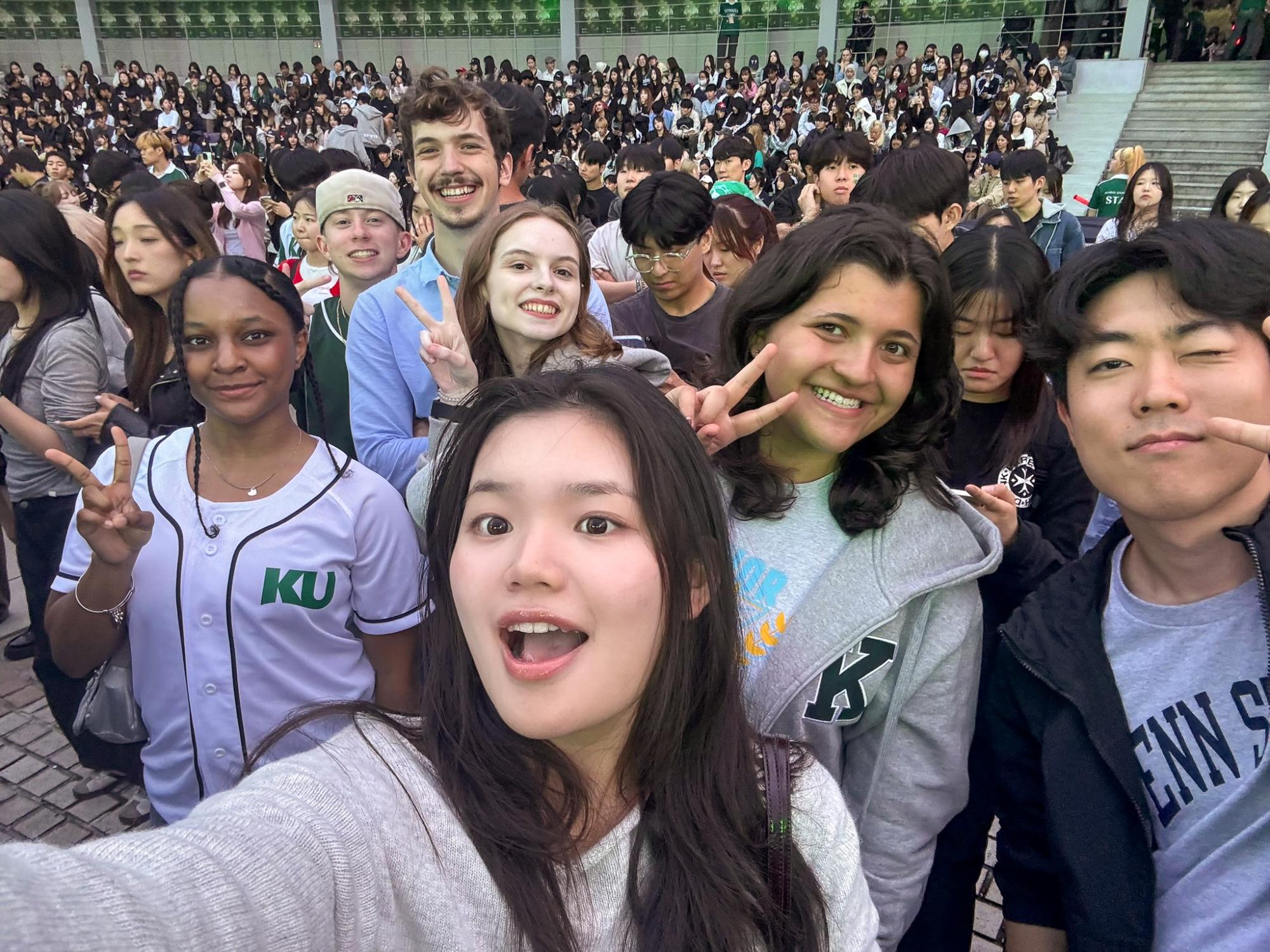 A group of people pose for a selfie in front of a large crowd, smiling and flashing peace signs.