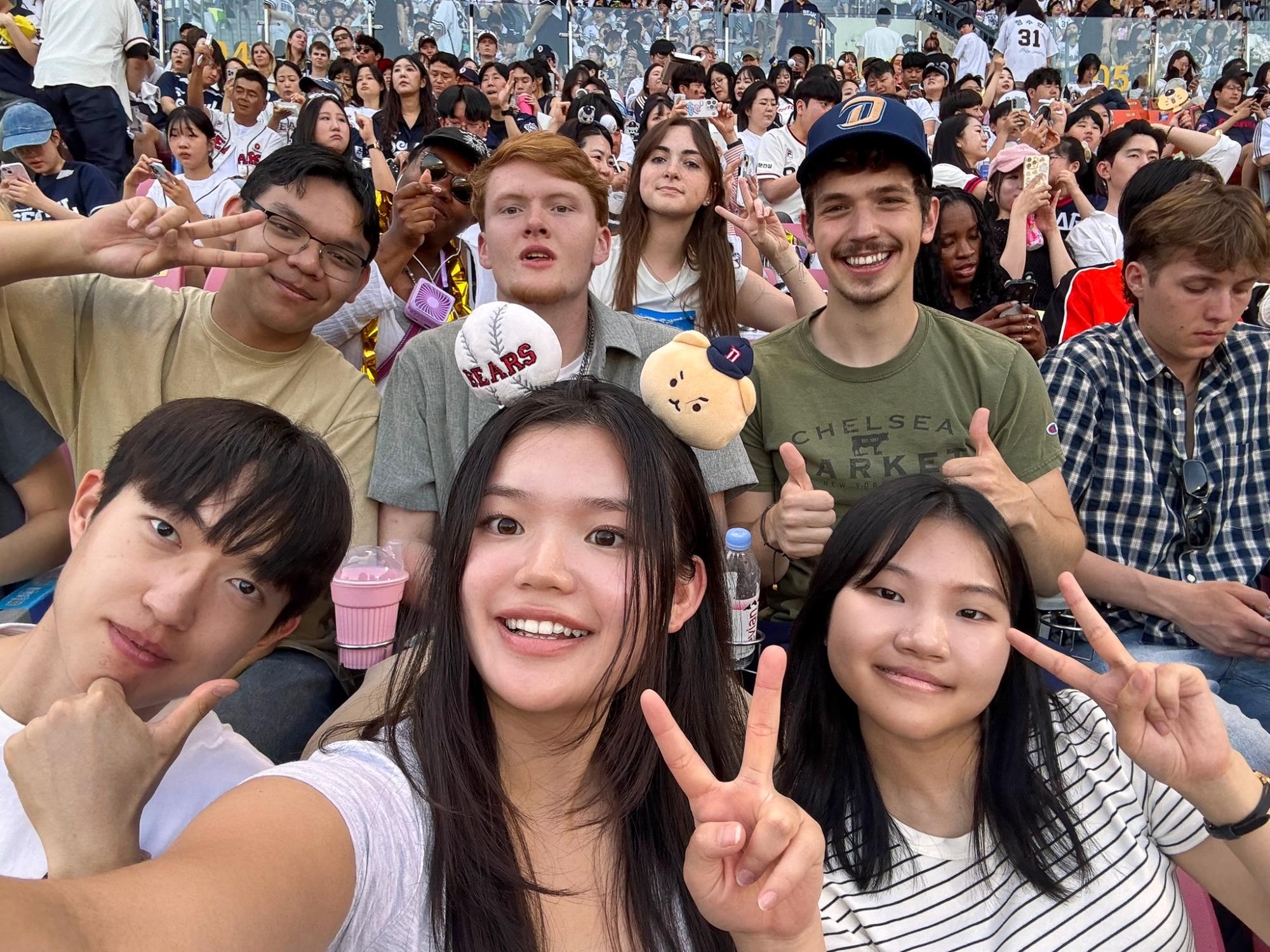 Six people pose for a selfie in a crowded baseball stadium filled with fans wearing jerseys and caps.
