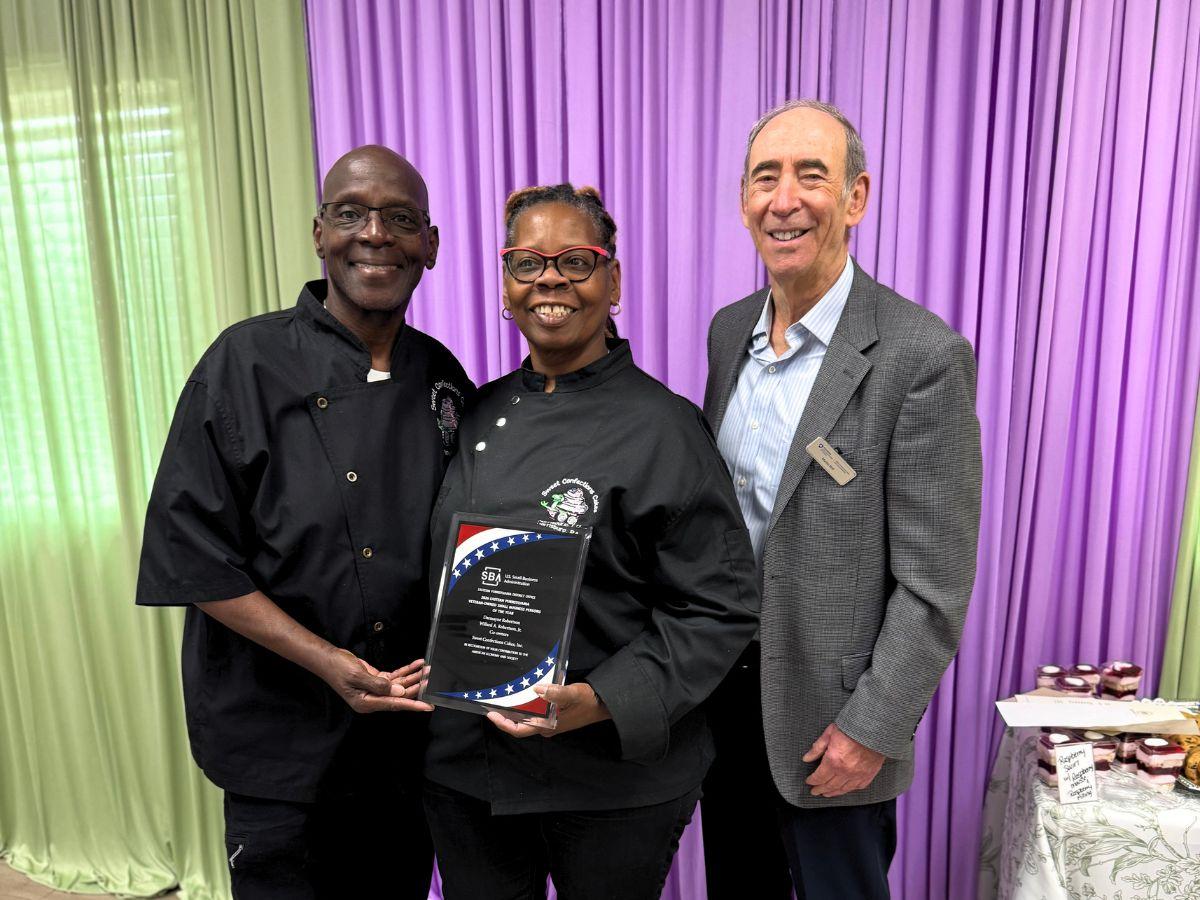 Three people pose together during an award presentation, with two chefs holding an SBA award plaque while standing beside a business representative in front of colorful draped backdrops.