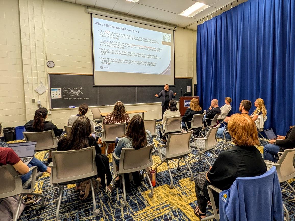 A man stands at the head of a room and gives a presentation.