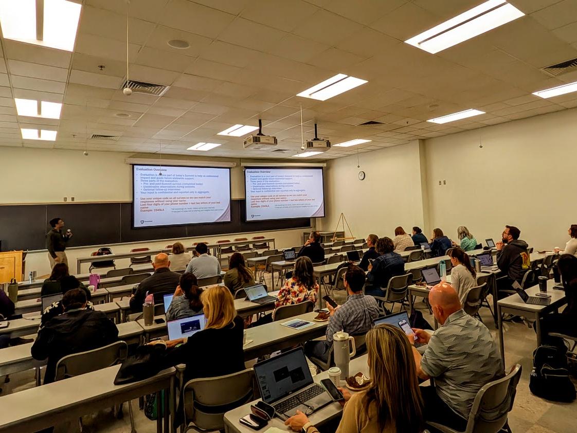A presenter stands in front of a seated crowd in a classroom