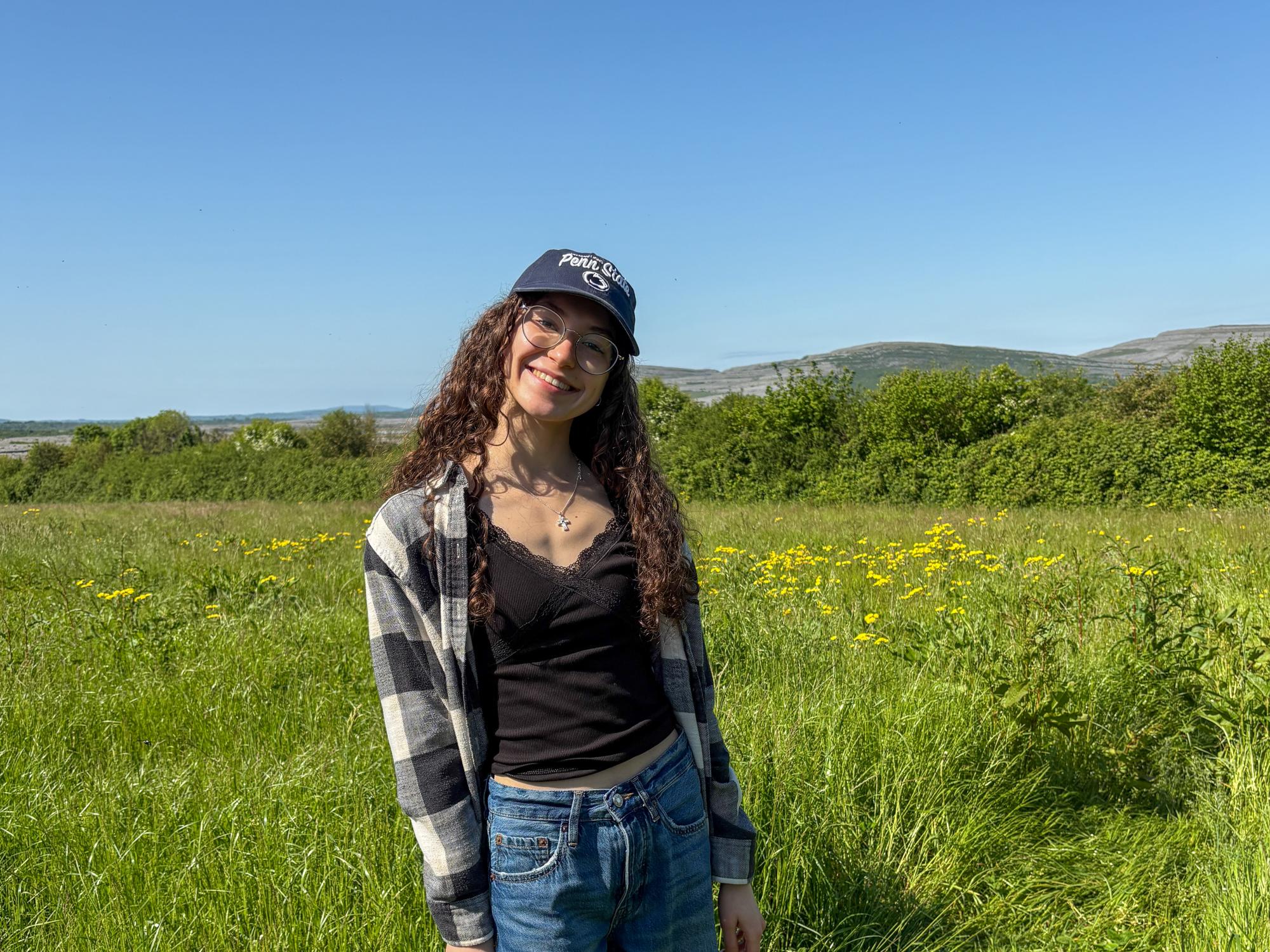 Chloe Smith, a young woman with curly hair, wearing a black tank top, plaid shirt and ripped jeans standing in a green field under a clear blue sky.