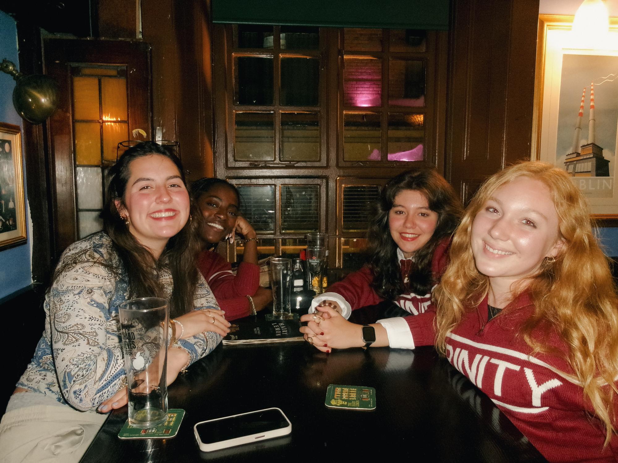 Four young women smiling and sitting around a dark wooden table in a cozy pub setting with drinks and a phone on the table.