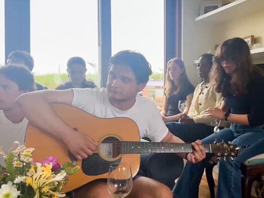 A group of students gathered indoors with one man playing an acoustic guitar and a woman playing the spoons while others listen attentively.