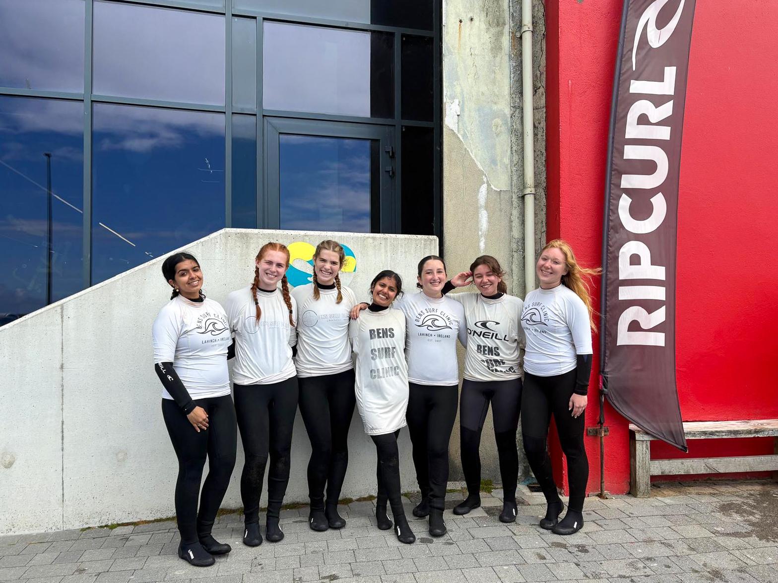 A group of seven young women in wetsuits standing together smiling outside a building with a Rip Curl banner nearby.