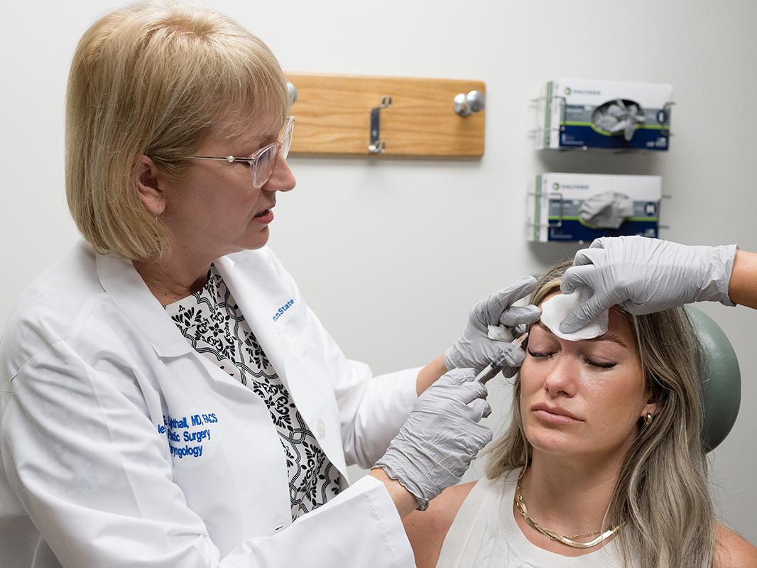 A physician injects Botox below the eyebrow of a woman patient. The hand of an assistant holds a piece of gauze on the patient’s forehead.