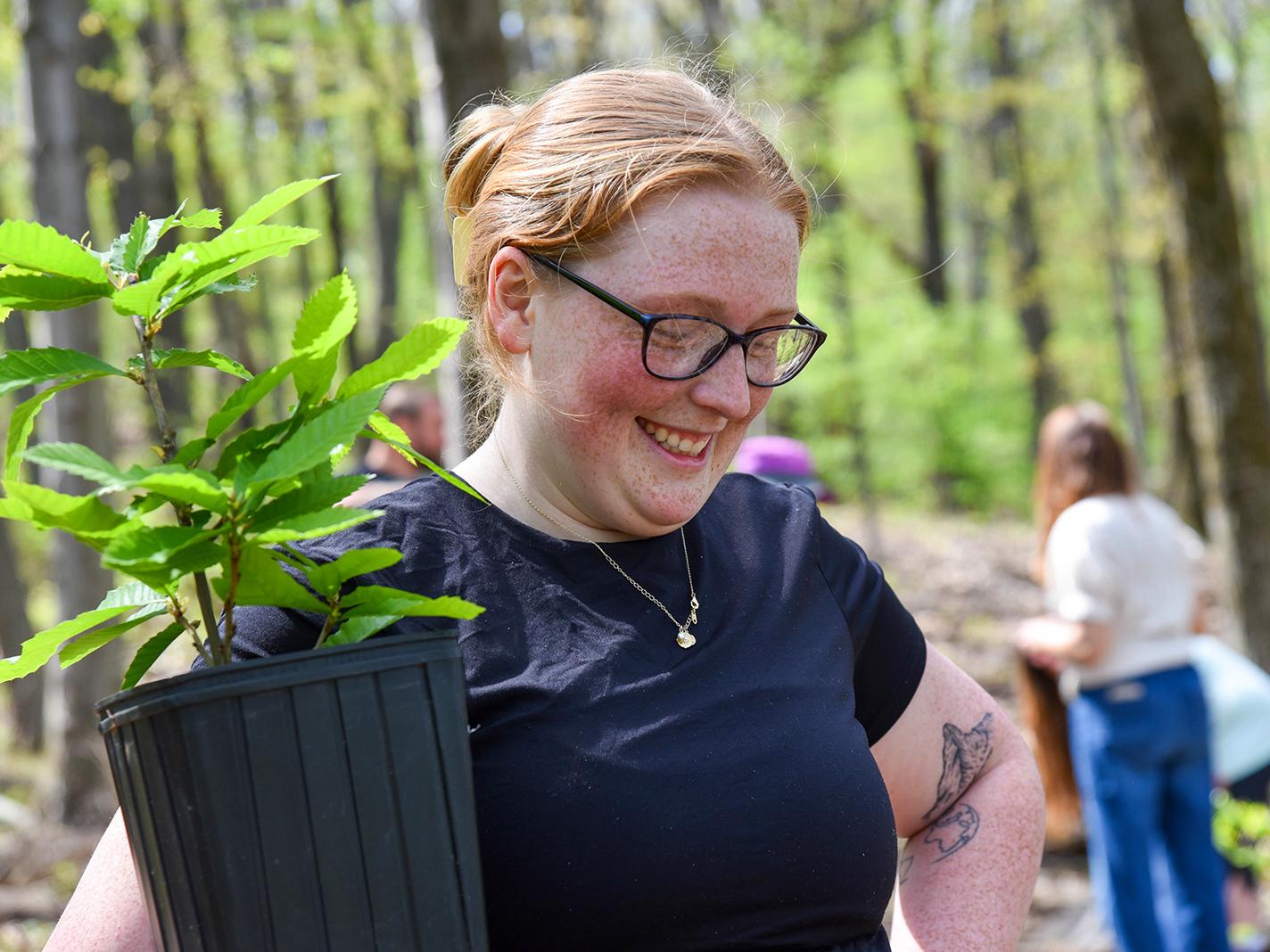 Falyn Verner smiles while holding a potted tree sapling in a wooded area.