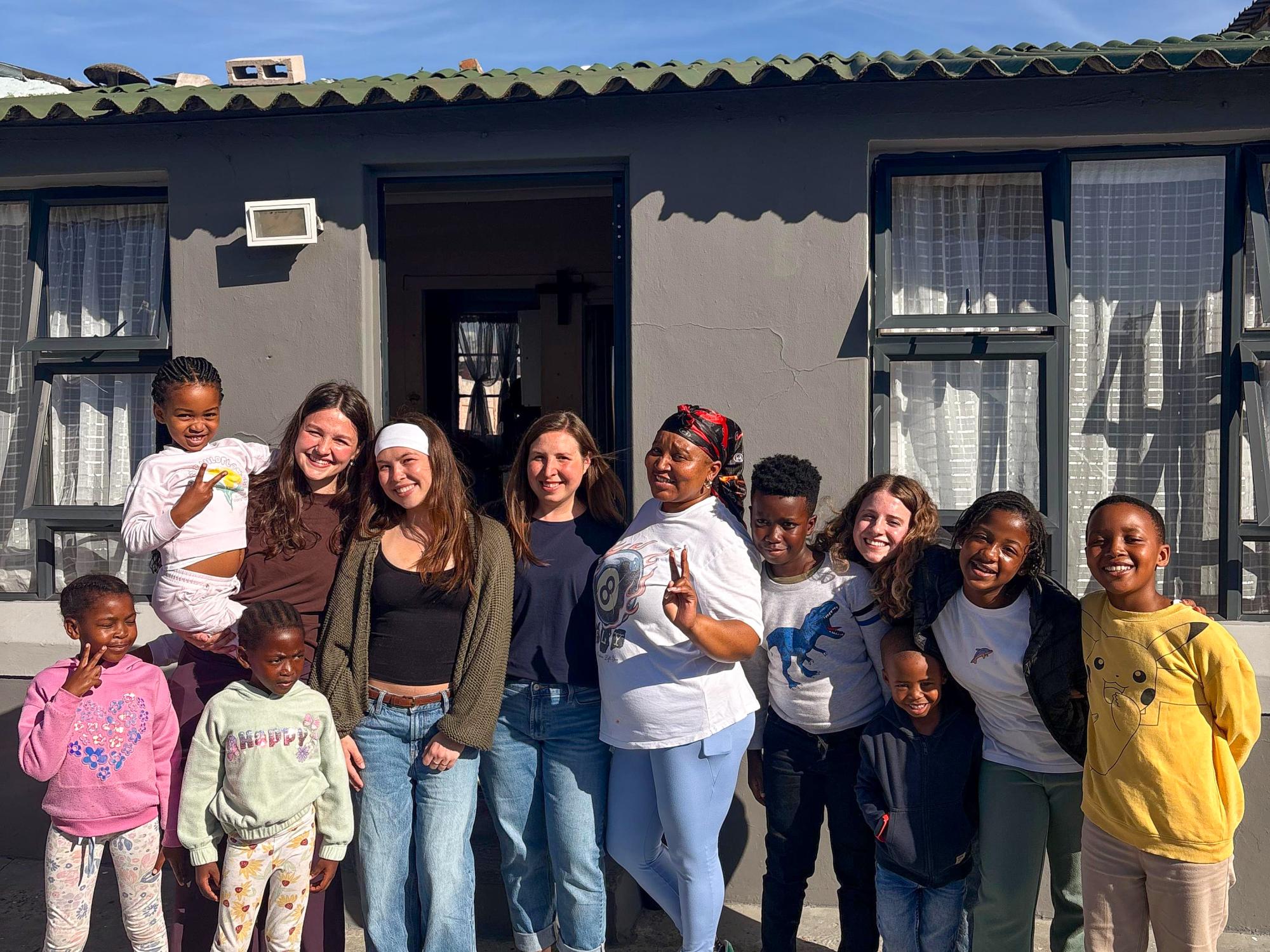 A diverse group of 12 people, including children and adults, are standing in front of a gray building with large windows. 