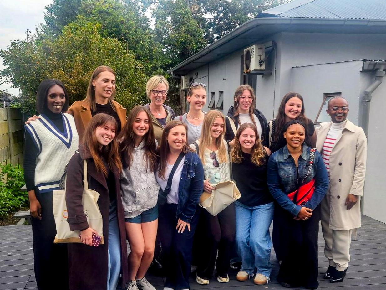 A diverse group of 13 individuals poses outdoors in front of a gray building, smiling and appearing friendly.