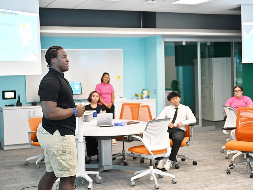 A high school student in a dark shirt gives a research presentation.