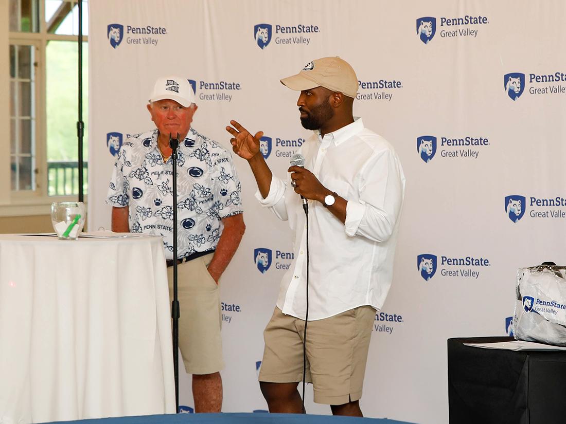 A man stands in front of Penn State Great Valley's backdrop and holds a microphone and speaks while another man watches him.