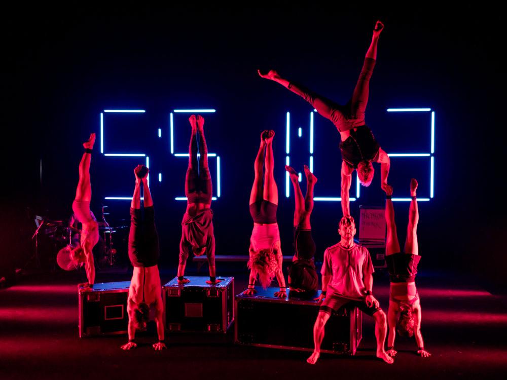 A group of athletes do handstands in front of a large digital time clock.