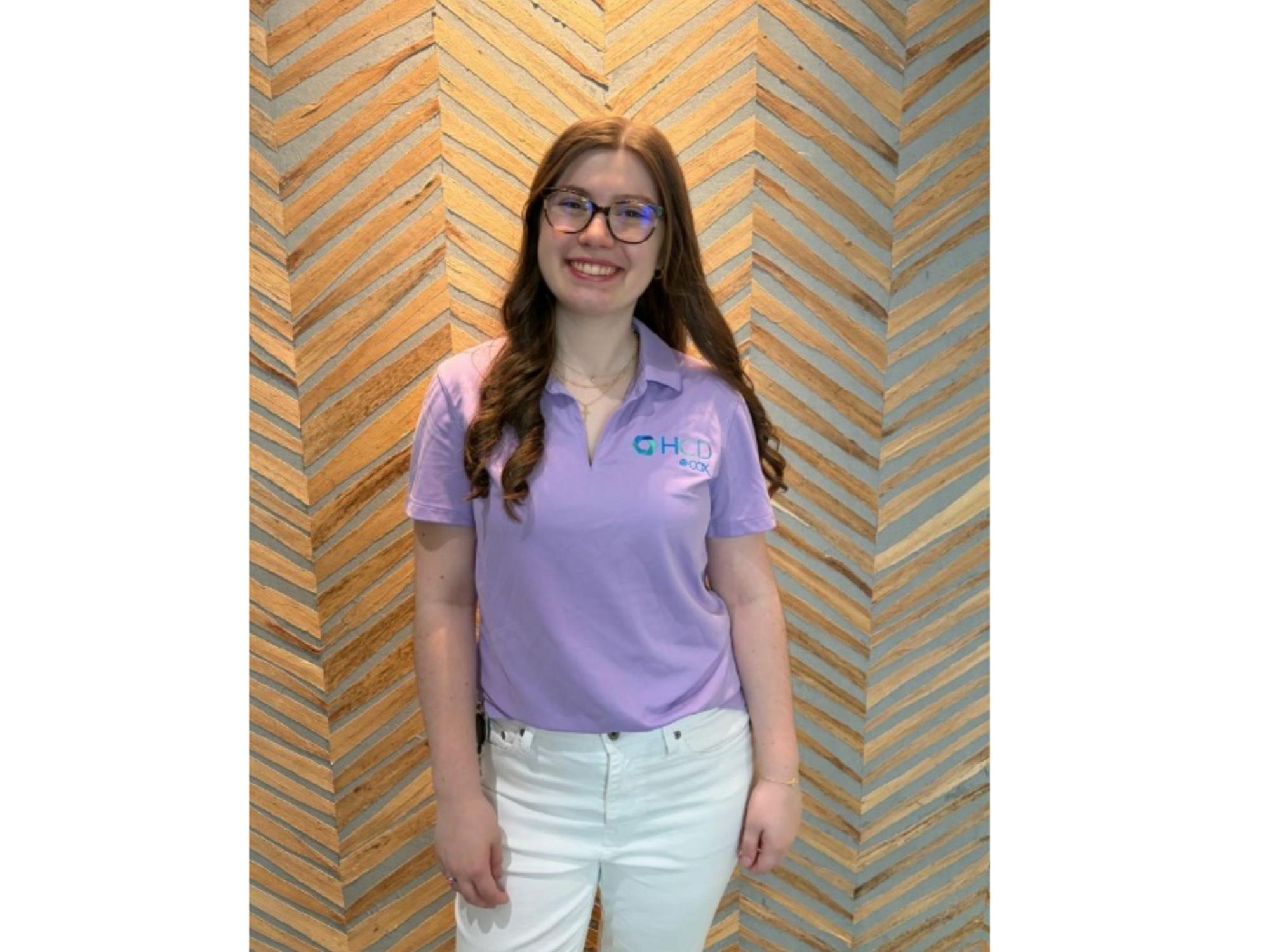 smiling person with glasses and long hair wearing purple shirt and white pants
