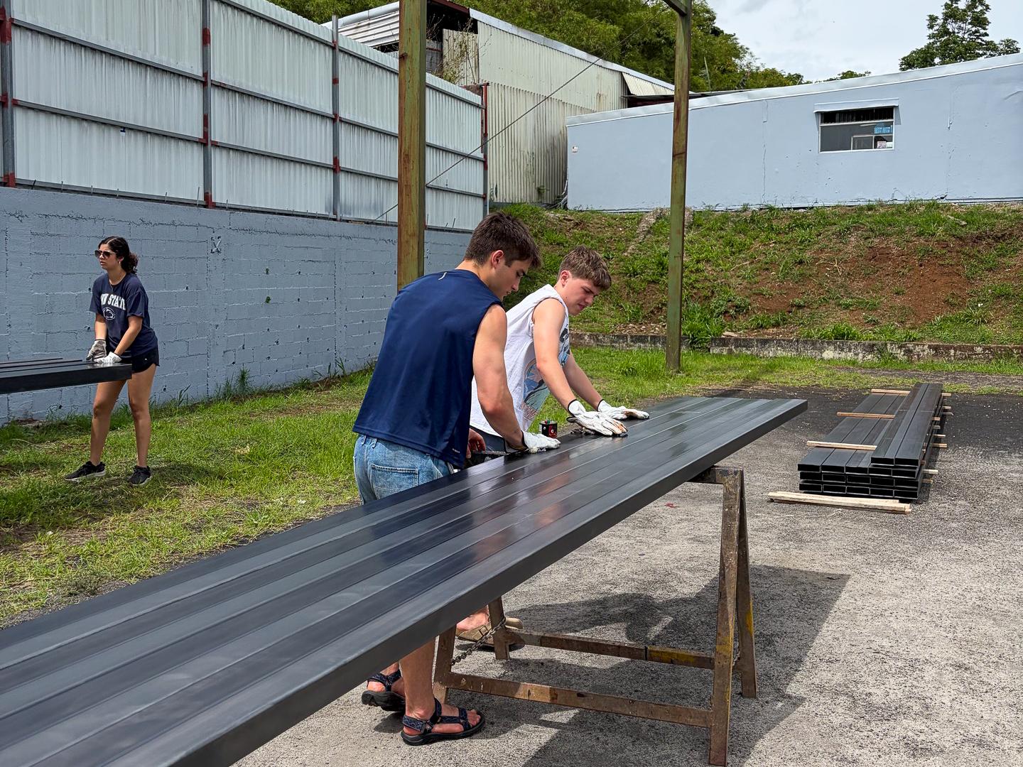 Students wearing gloves prepare and paint beams in an outdoor space.