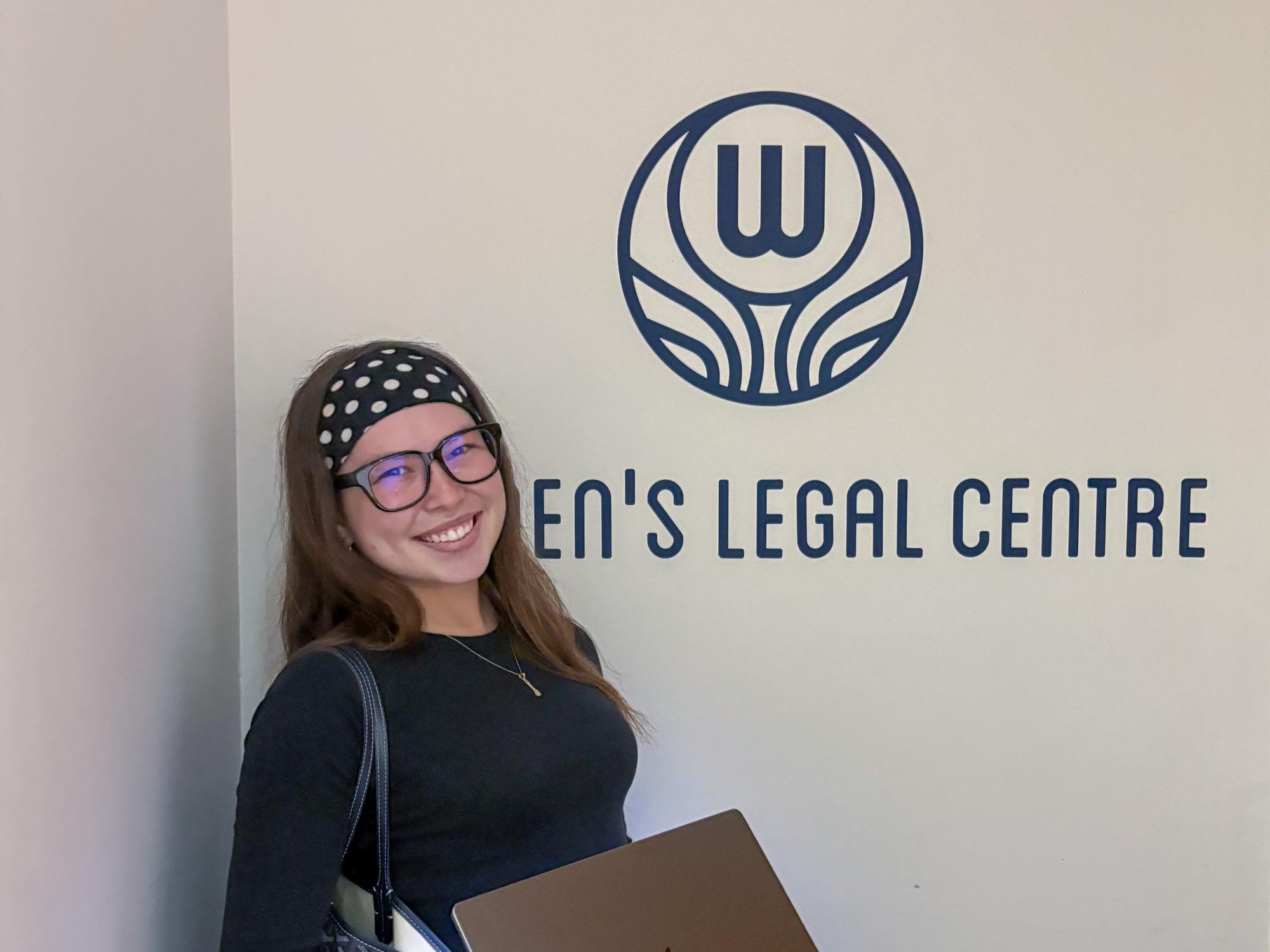 A young woman with long brown hair and glasses stands beside a wall that reads “Women’s Legal Centre,” holding a laptop and a patterned tote bag.