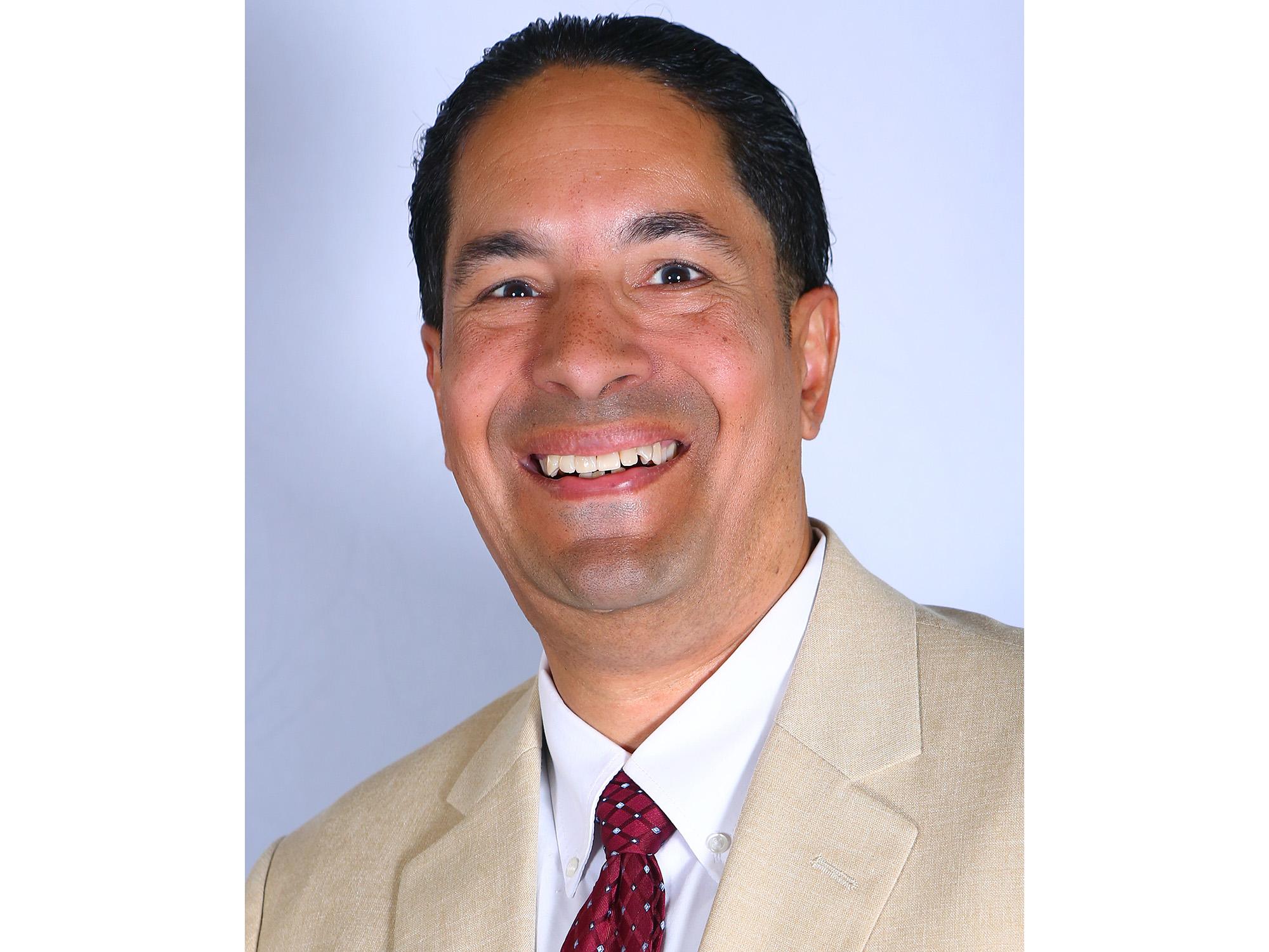 Michael Toledo, smiling and wearing a tan suit jacket and burgundy tie