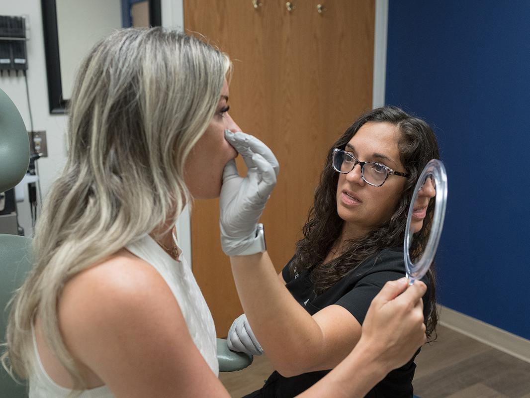 A facial nerve therapist performs therapy on a woman by gently positioning her facial muscles. The patient holds a hand mirror while watching the treatment.