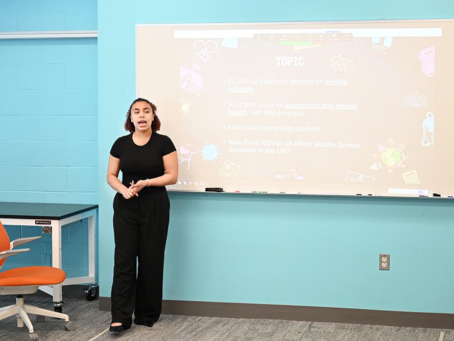 A high school student in a dark outfit gives a research presentation.