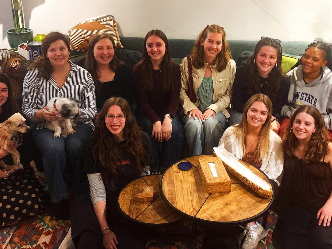 A diverse group of 10 people, and two dogs sit on a green couch and around a round coffee table. 