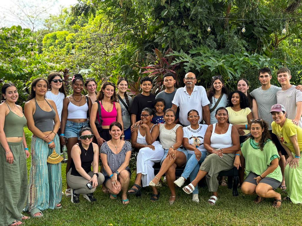 A group of students, staff, and a family pose together outdoors surrounded by greenery.