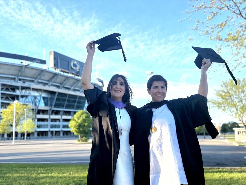 Two graduates in academic regalia stand outdoors near a stadium, holding graduation caps overhead on a sunny day.