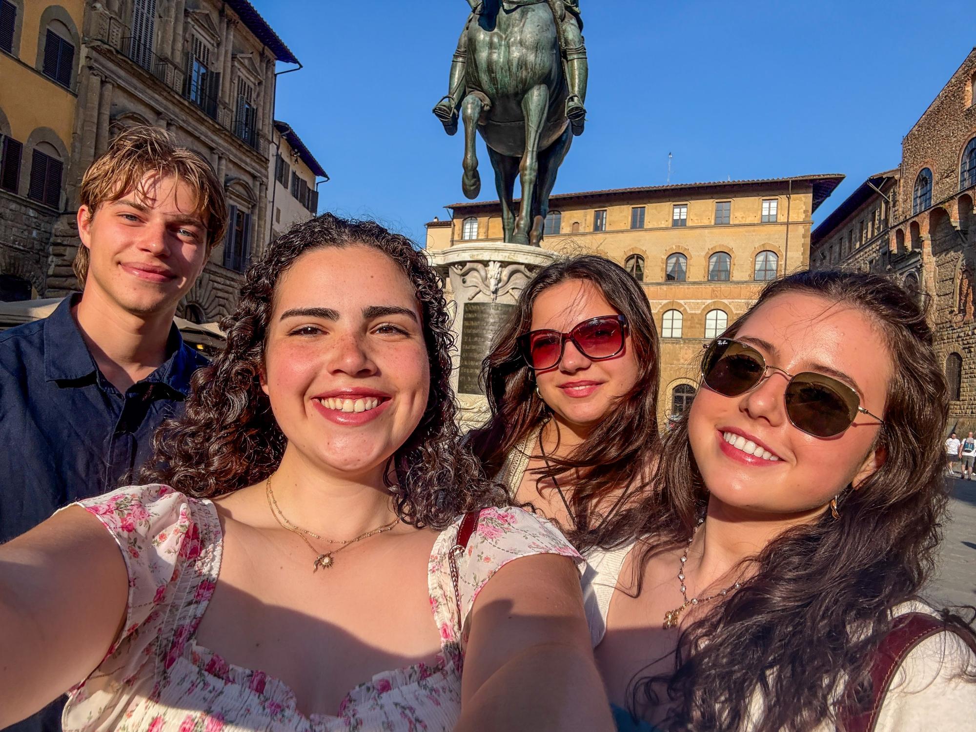 Four people smile for a selfie in front of a large equestrian statue in a historic European square, surrounded by stone buildings and bright blue sky.