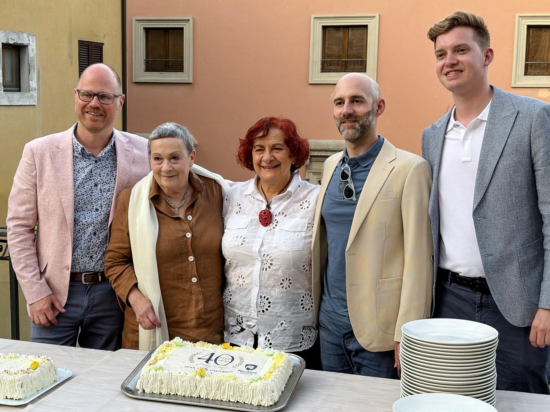 Five people stand behind a table with two decorated cakes which feature a “40” emblem and laurel wreath set in an outdoor courtyard.