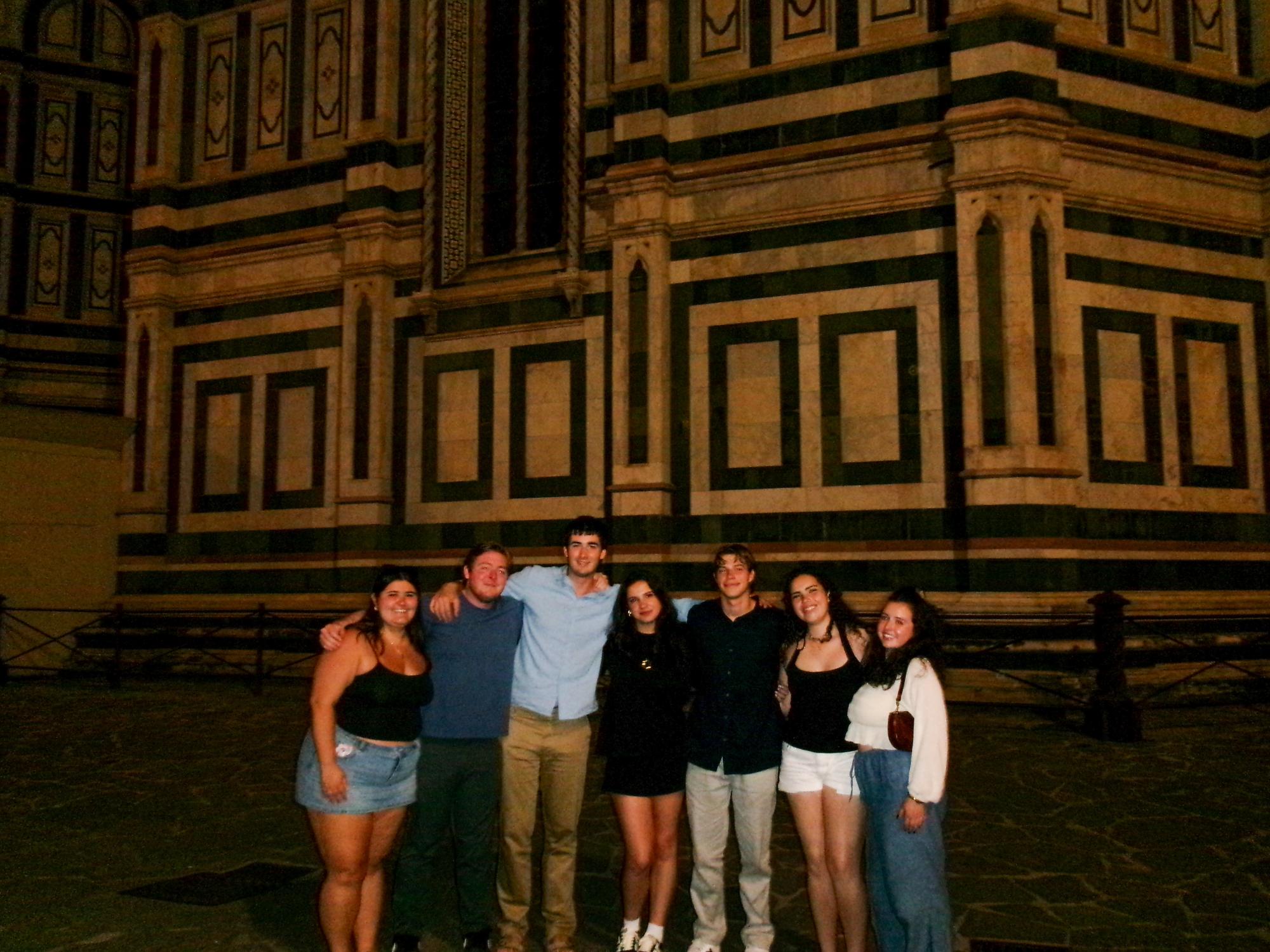 Seven people stand together at night in front of a large marble-patterned historic building featuring geometric designs.