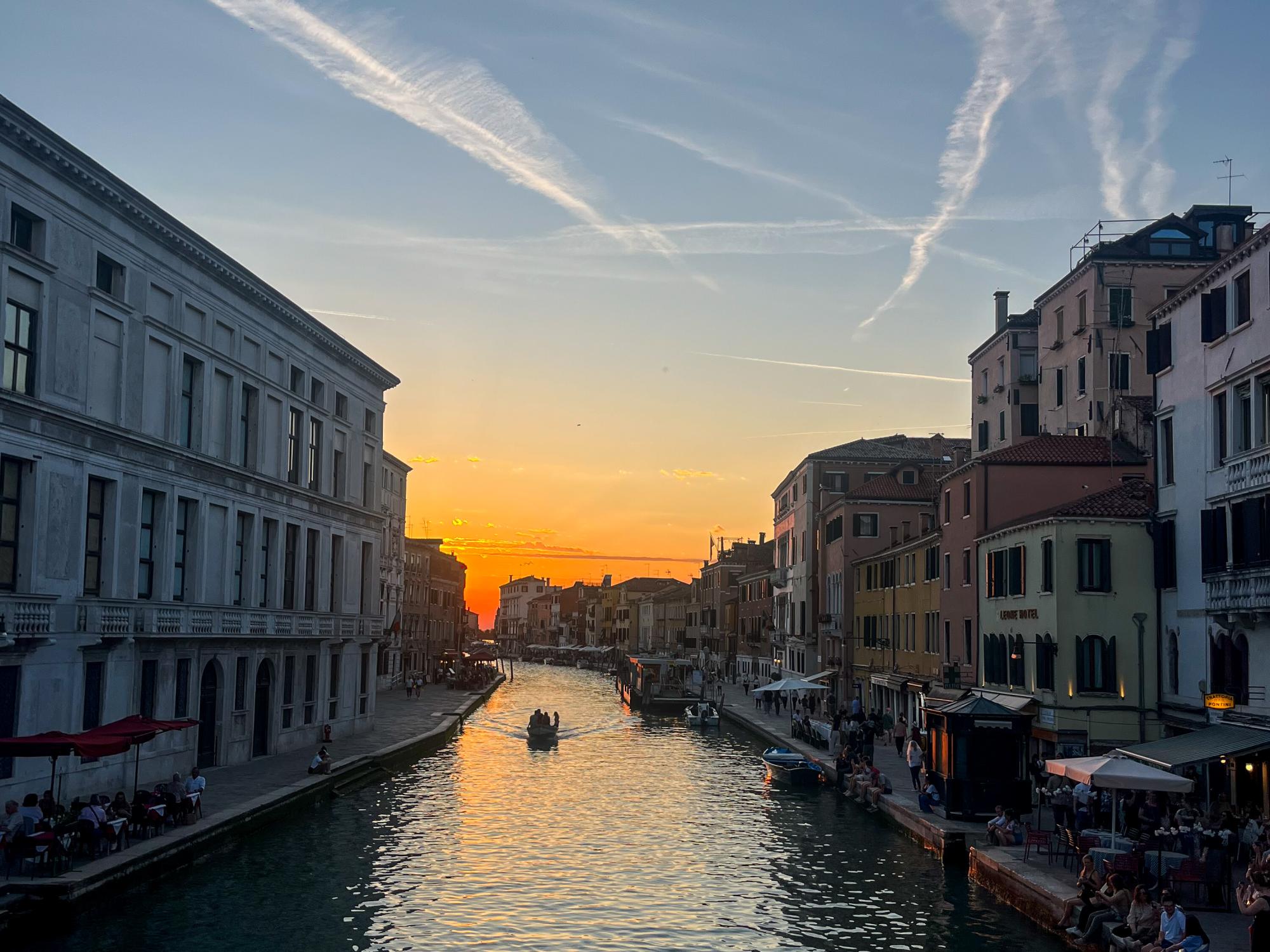 A sunset casts warm light over a Venetian canal lined with historic buildings and a small boat moving through the water.