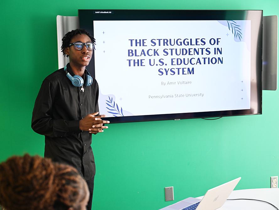 A high school student wearing a dark outfit gives a research presentation.