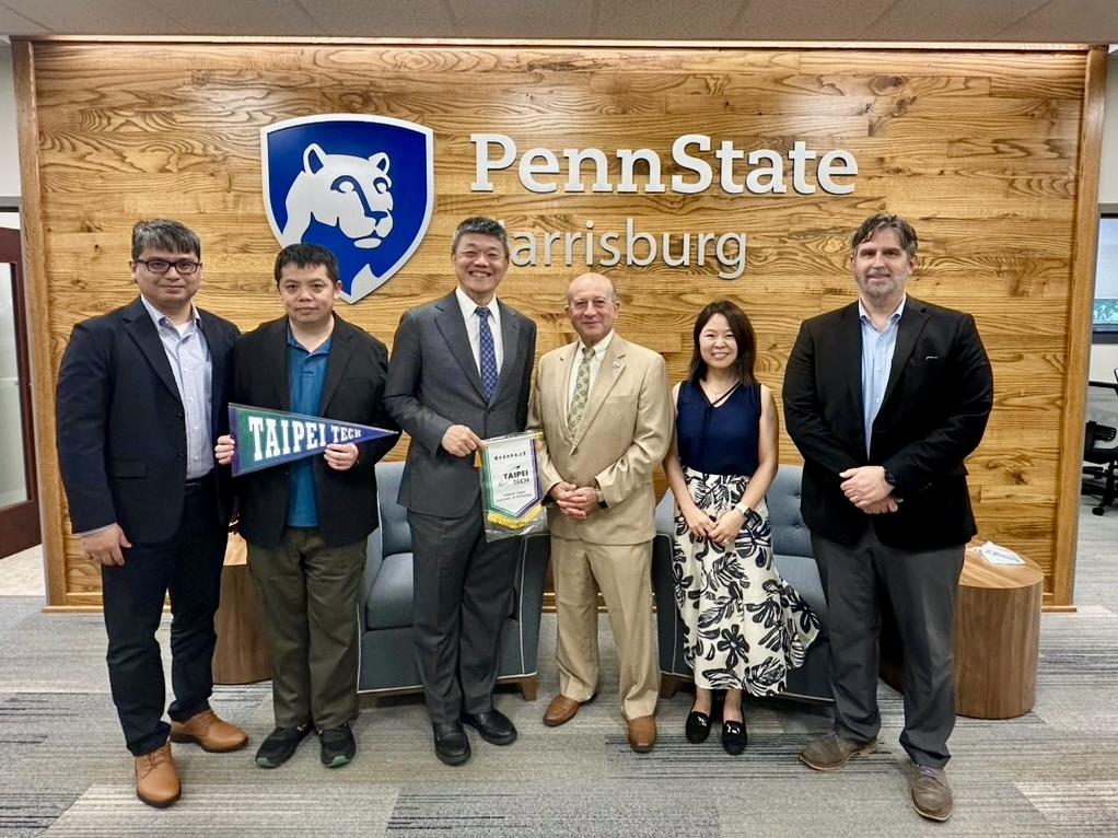 A group smiles in front of a Penn State Harrisburg sign; one of them is holding a Taipei Tech pennant.