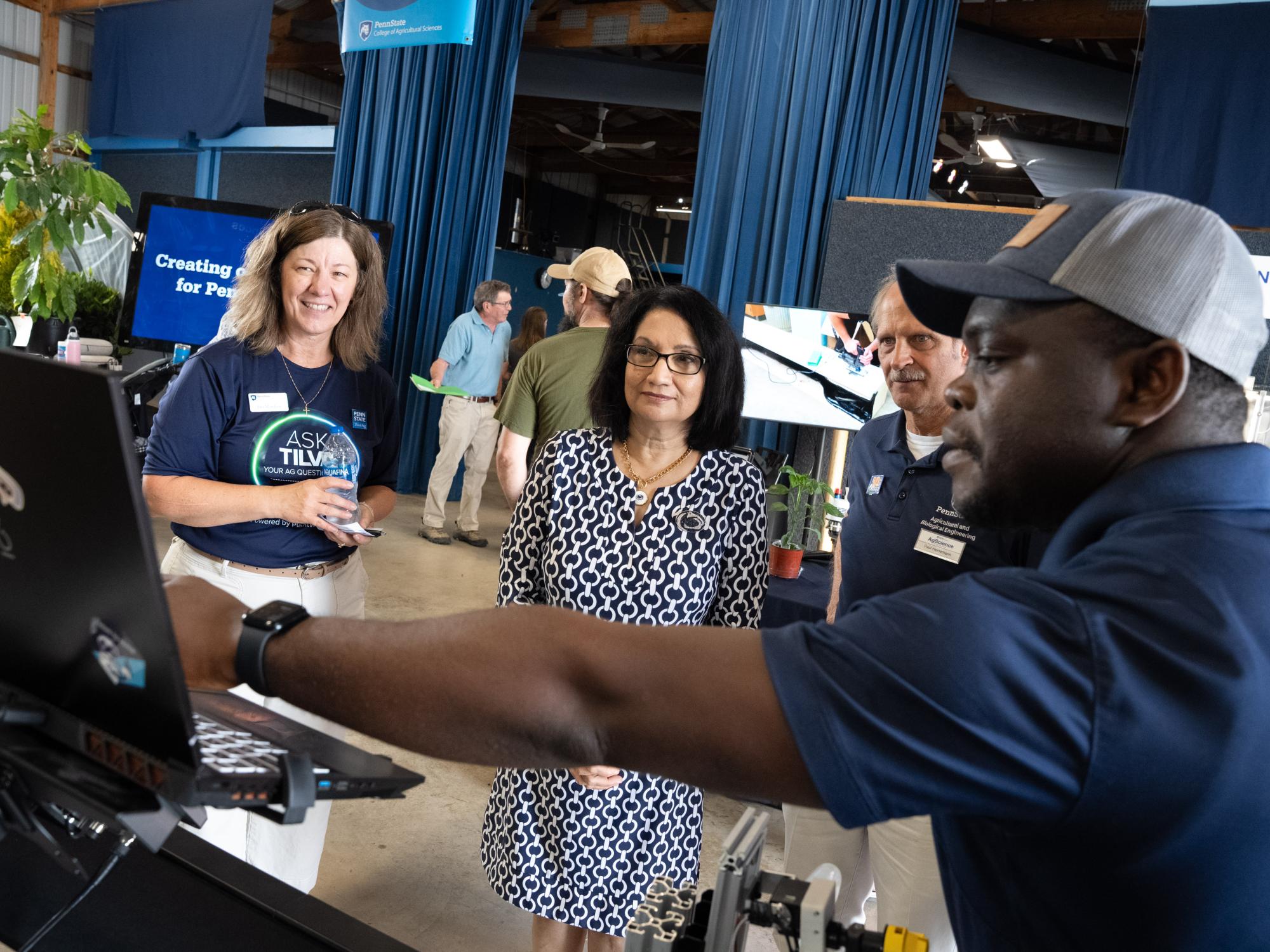 A person explains information displayed on a laptop screen to three visitors at a booth