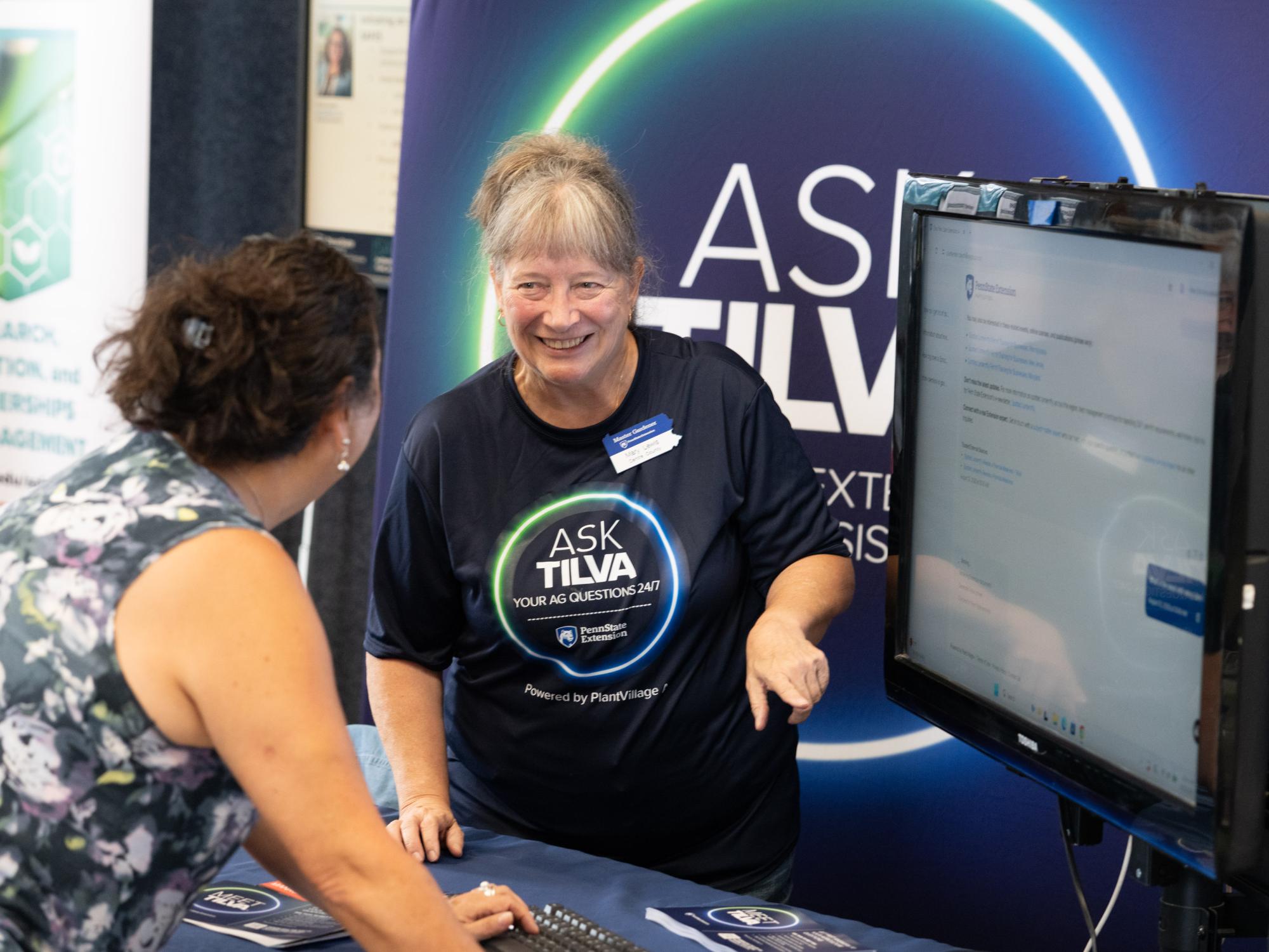 A person wearing a shirt that reads "Ask Tilva" explains information on a computer screen to a booth visitor