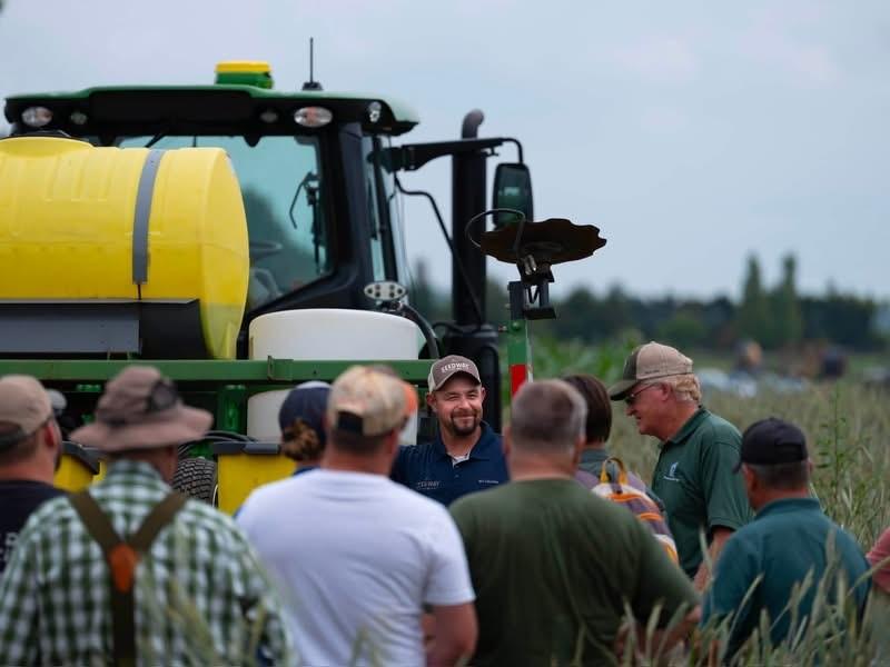 A small group of individuals with their backs to the camera listen to a man standing next to a tractor