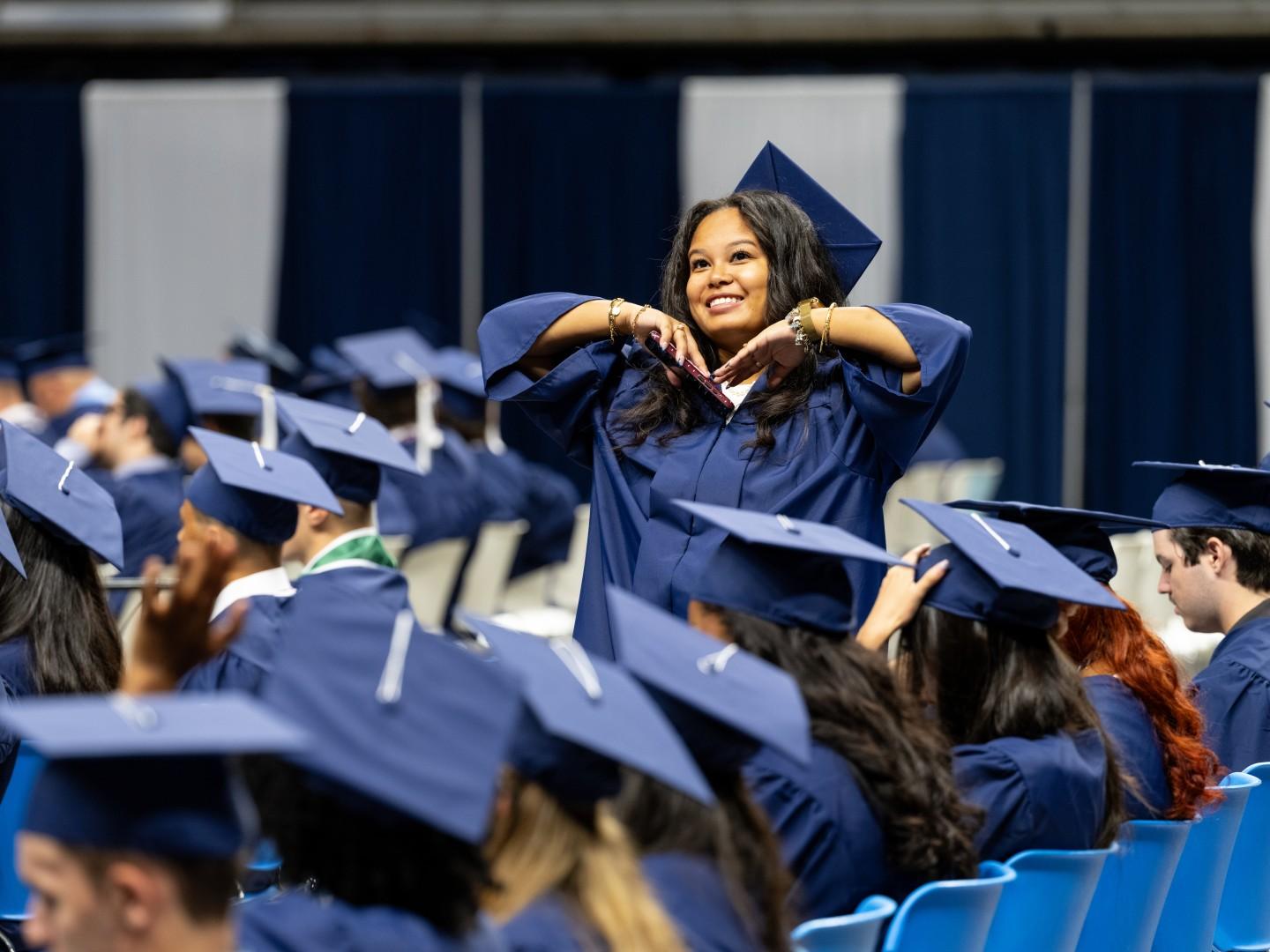 A graduate wearing a blue cap and gown stands among a crowd of seated peers