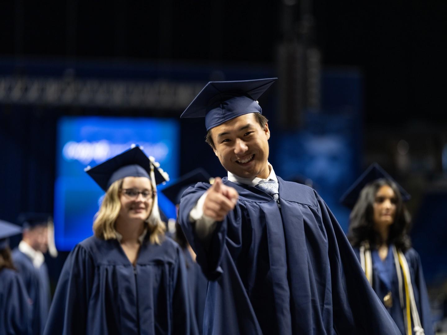 A person wearing a blue cap and gown smiles and points at the camera